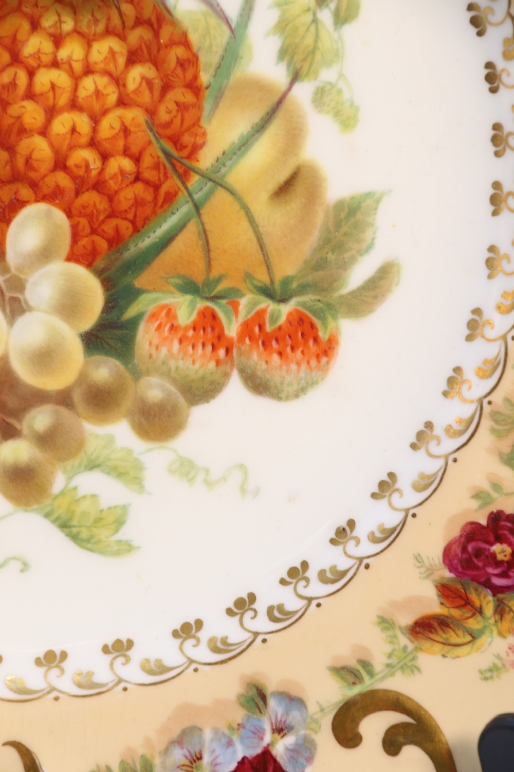 A cream-coloured circular porcelain cabinet plate featuring a scalloped gilded rim, floral swags, and a central hand-painted motif of a pineapple, grapes, and fruit. View XVIII