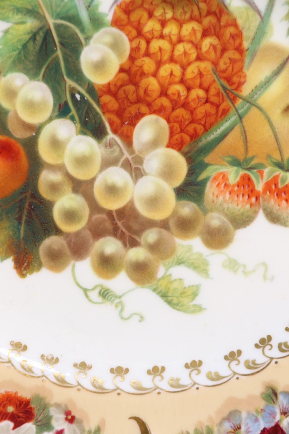 A cream-coloured circular porcelain cabinet plate featuring a scalloped gilded rim, floral swags, and a central hand-painted motif of a pineapple, grapes, and fruit. View XIX