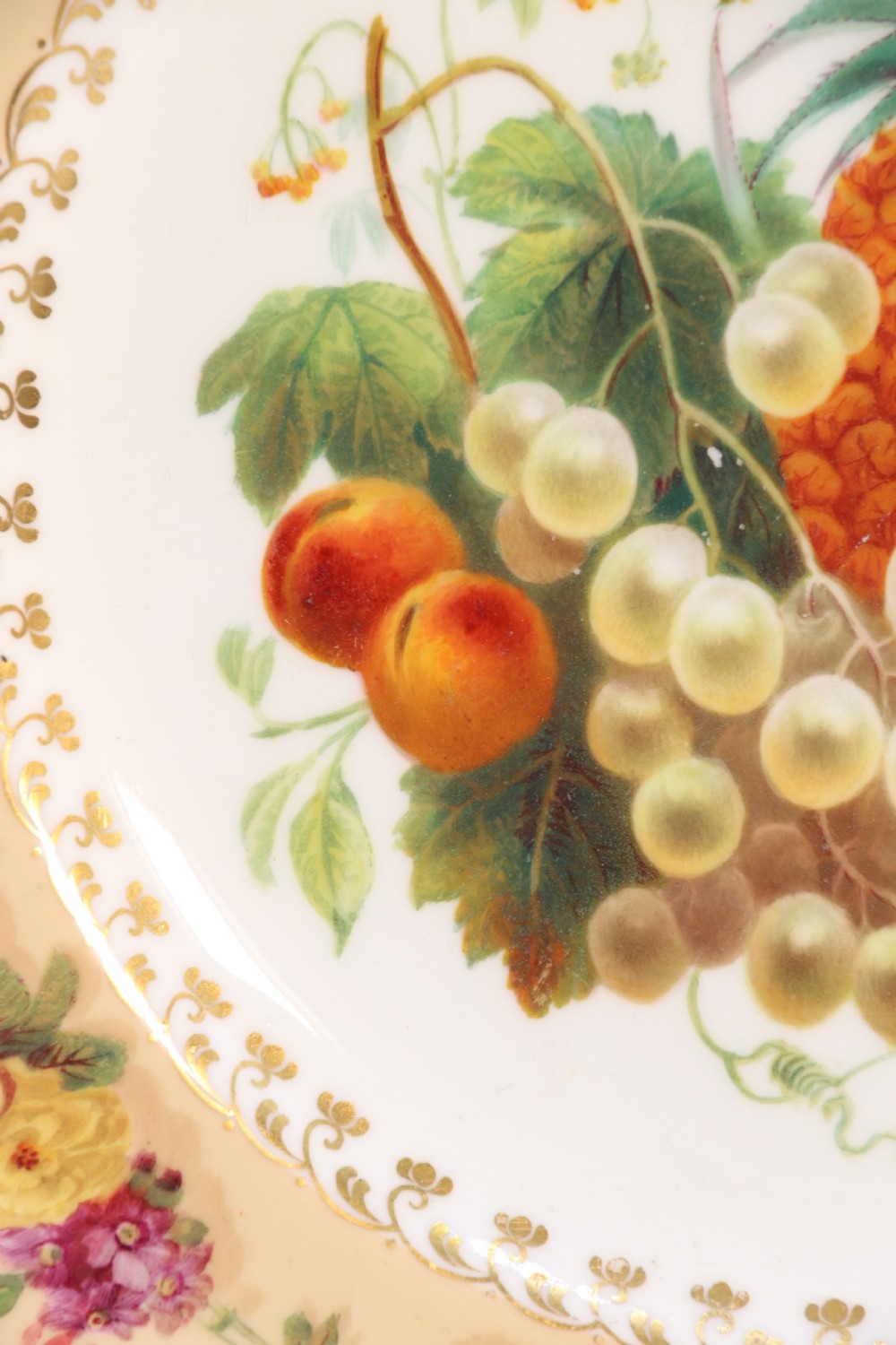 A cream-coloured circular porcelain cabinet plate featuring a scalloped gilded rim, floral swags, and a central hand-painted motif of a pineapple, grapes, and fruit. View XX