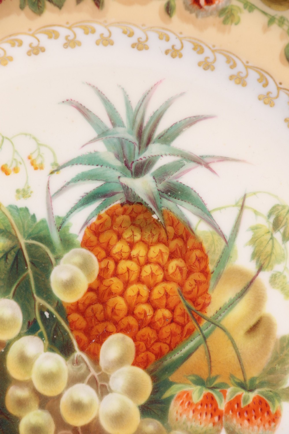 A cream-coloured circular porcelain cabinet plate featuring a scalloped gilded rim, floral swags, and a central hand-painted motif of a pineapple, grapes, and fruit. View XXI