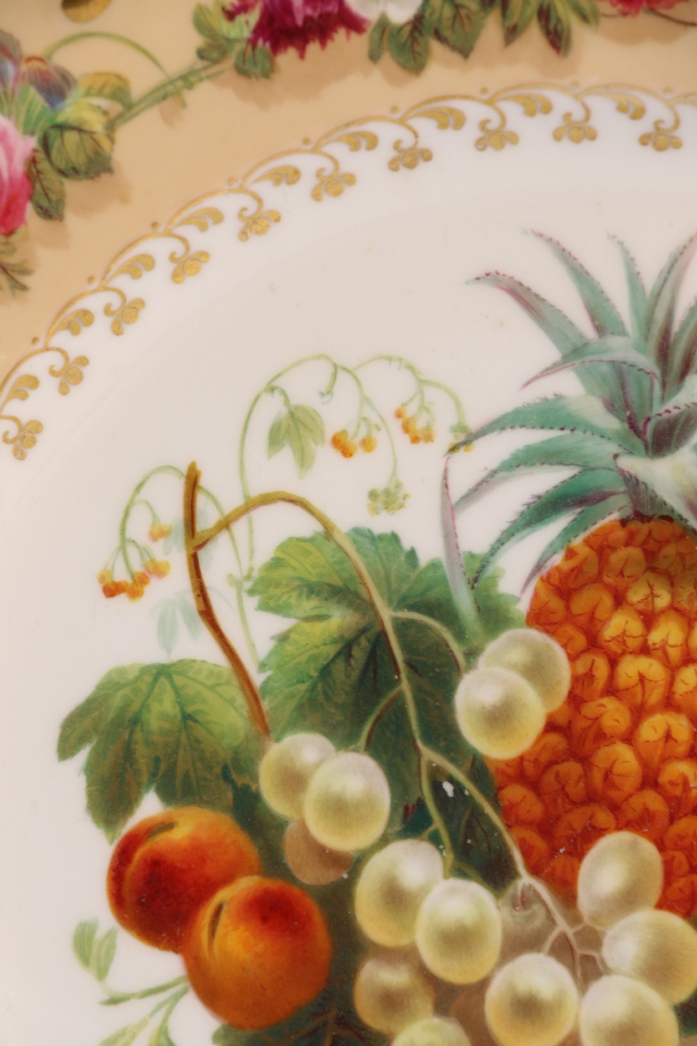 A cream-coloured circular porcelain cabinet plate featuring a scalloped gilded rim, floral swags, and a central hand-painted motif of a pineapple, grapes, and fruit. View XXII