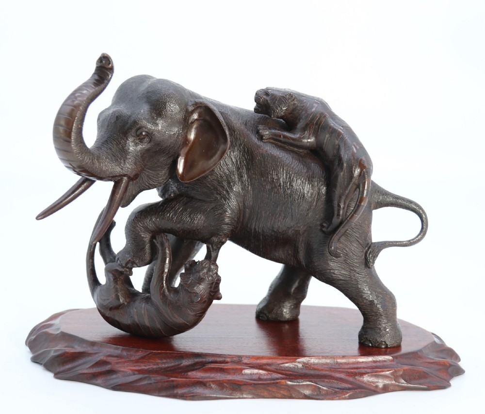 A dark patinated bronze sculpture depicting an elephant defending itself against two attacking tigers, mounted on a contoured wooden base.