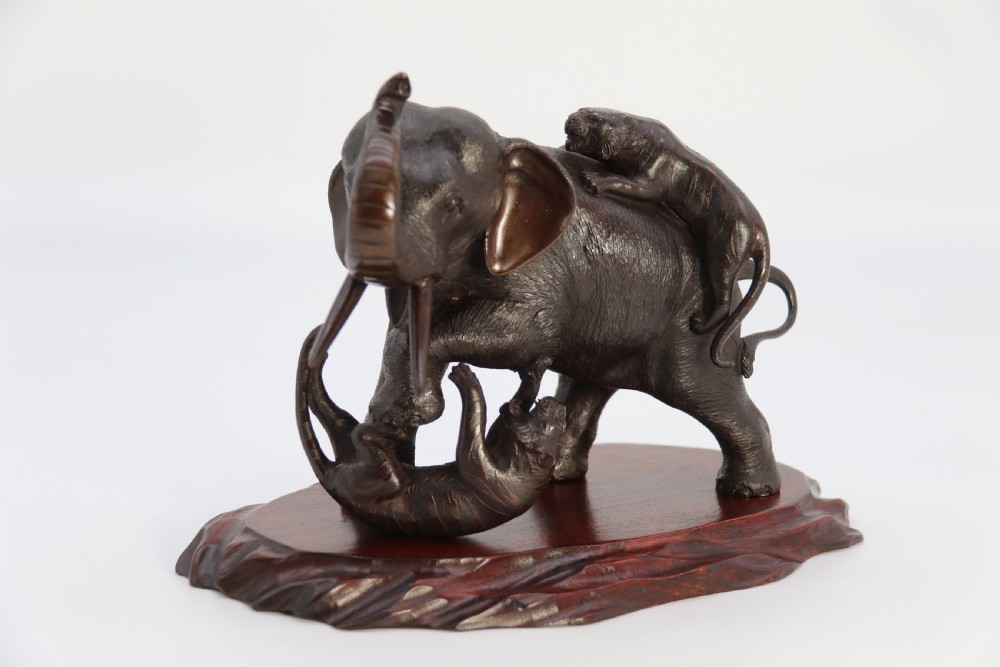 A dark patinated bronze sculpture depicting an elephant defending itself against two attacking tigers, mounted on a contoured wooden base. View II