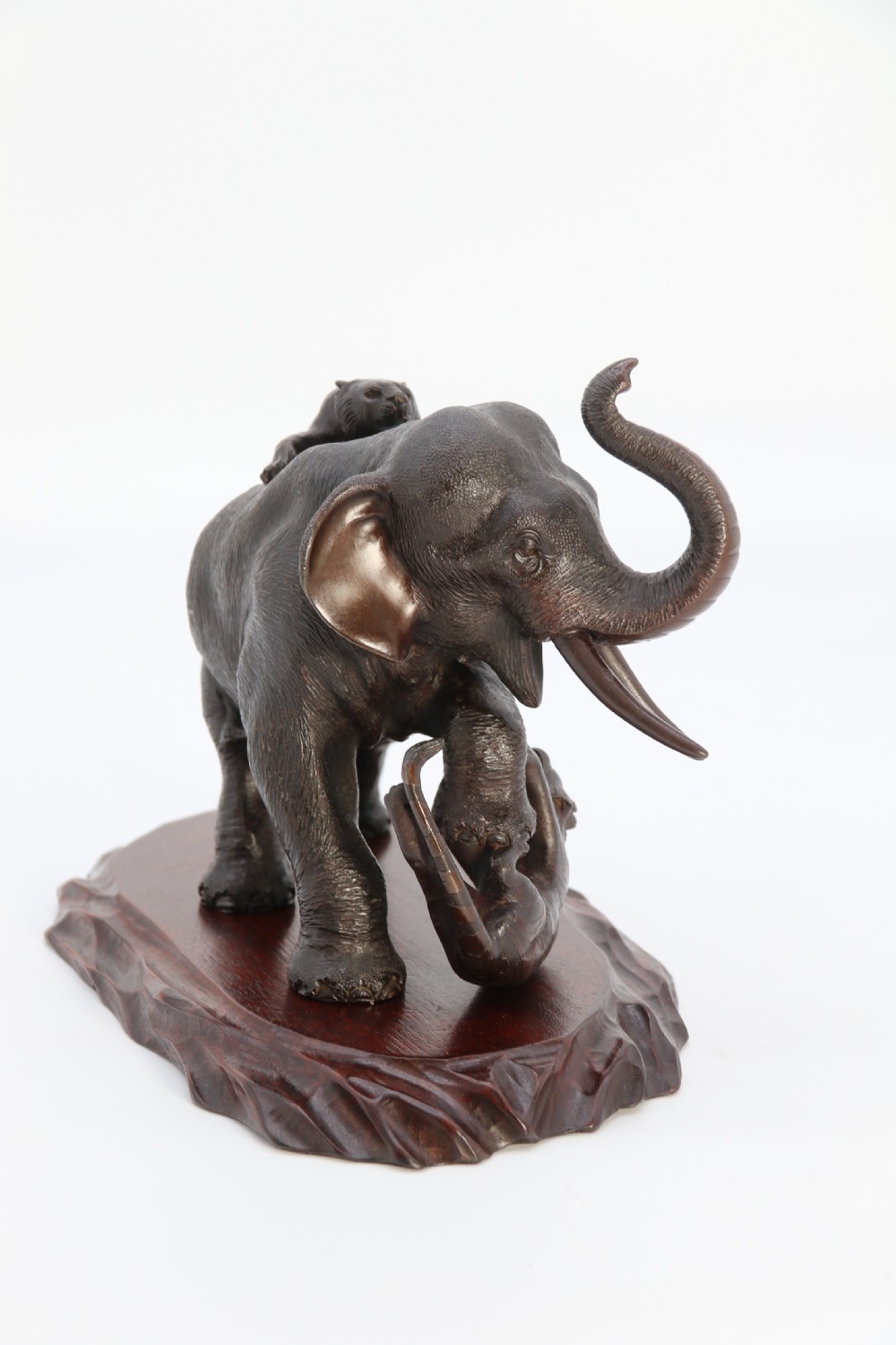A dark patinated bronze sculpture depicting an elephant defending itself against two attacking tigers, mounted on a contoured wooden base. View III