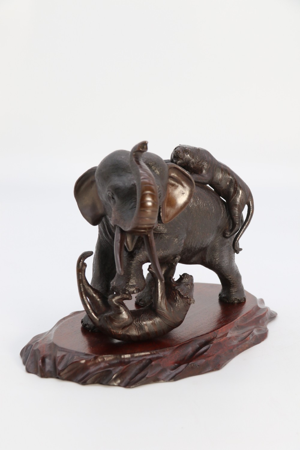 A dark patinated bronze sculpture depicting an elephant defending itself against two attacking tigers, mounted on a contoured wooden base. View IV