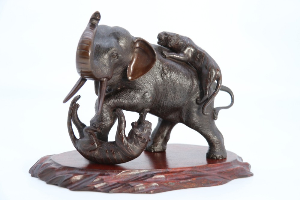 A dark patinated bronze sculpture depicting an elephant defending itself against two attacking tigers, mounted on a contoured wooden base. View V