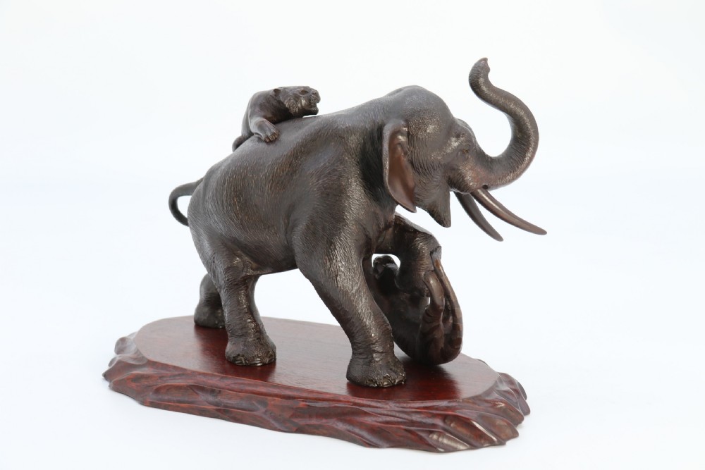 A dark patinated bronze sculpture depicting an elephant defending itself against two attacking tigers, mounted on a contoured wooden base. View IX