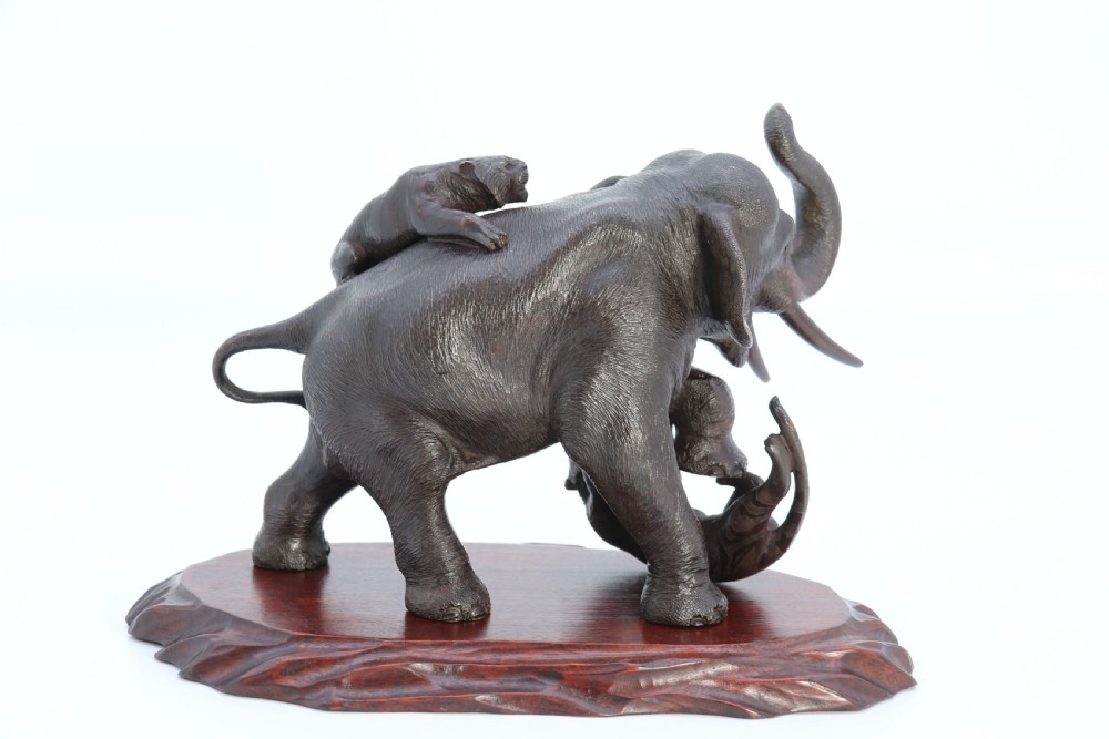 A dark patinated bronze sculpture depicting an elephant defending itself against two attacking tigers, mounted on a contoured wooden base. View X