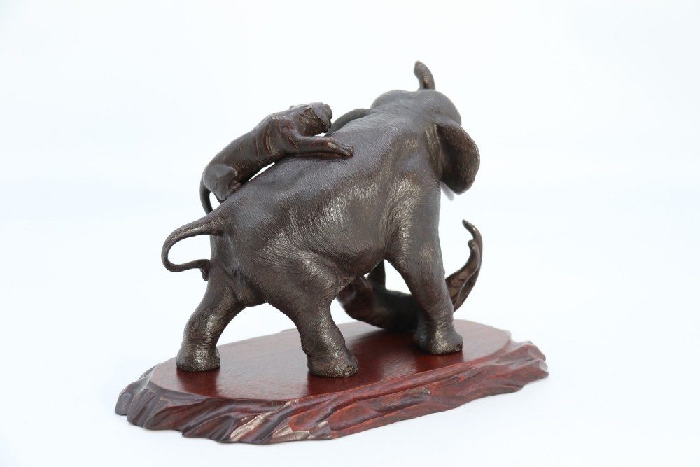 A dark patinated bronze sculpture depicting an elephant defending itself against two attacking tigers, mounted on a contoured wooden base. View XI