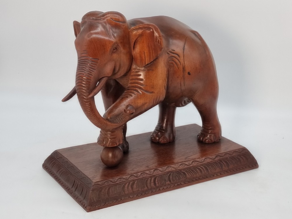 A pair of hand-carved warm brown teak wooden elephant sculptures balancing on balls mounted on rectangular bases. View II