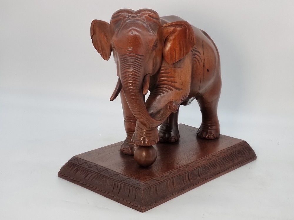 A pair of hand-carved warm brown teak wooden elephant sculptures balancing on balls mounted on rectangular bases. View III