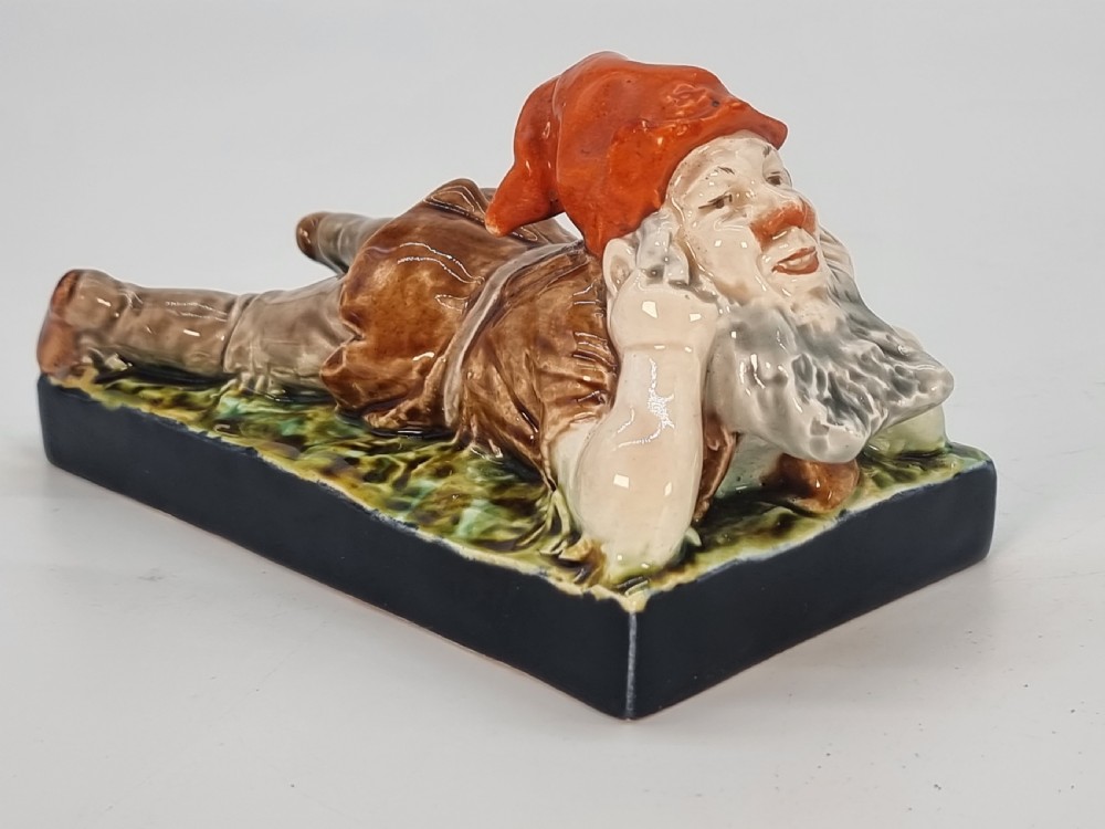 A Victorian majolica glazed earthenware paperweight depicting a reclining gnome with a red cap, grey beard, and brown tunic on a dark rectangular base.