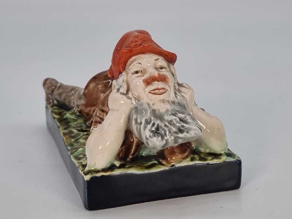 A Victorian majolica glazed earthenware paperweight depicting a reclining gnome with a red cap, grey beard, and brown tunic on a dark rectangular base. View II