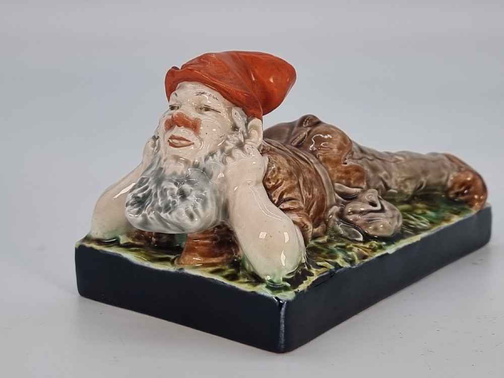 A Victorian majolica glazed earthenware paperweight depicting a reclining gnome with a red cap, grey beard, and brown tunic on a dark rectangular base. View III