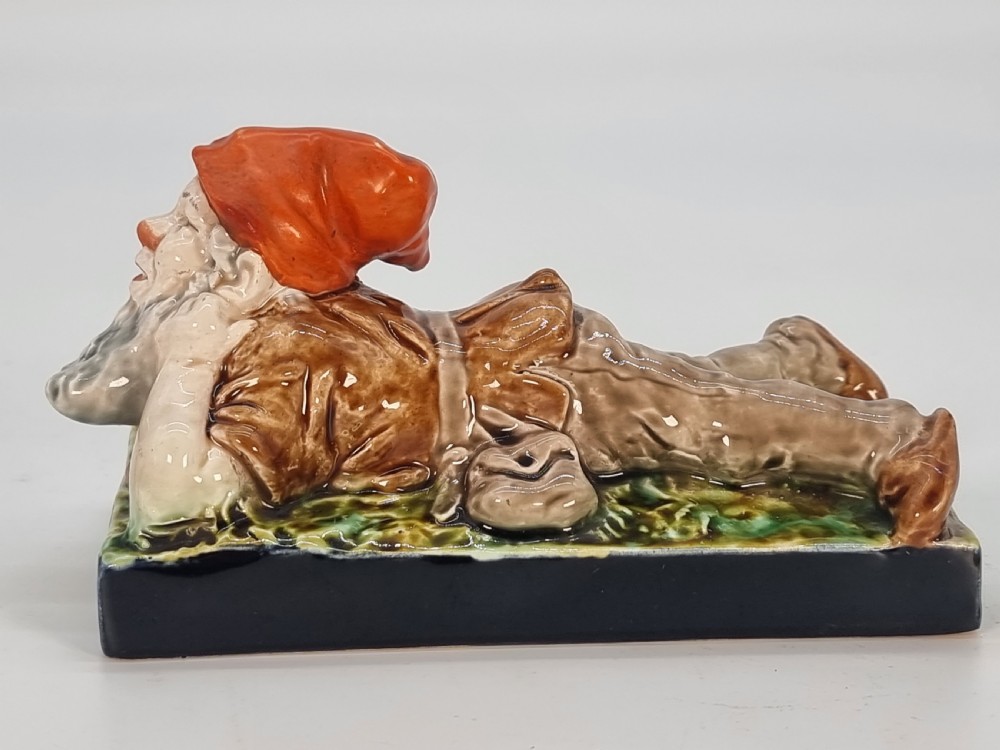 A Victorian majolica glazed earthenware paperweight depicting a reclining gnome with a red cap, grey beard, and brown tunic on a dark rectangular base. View IV
