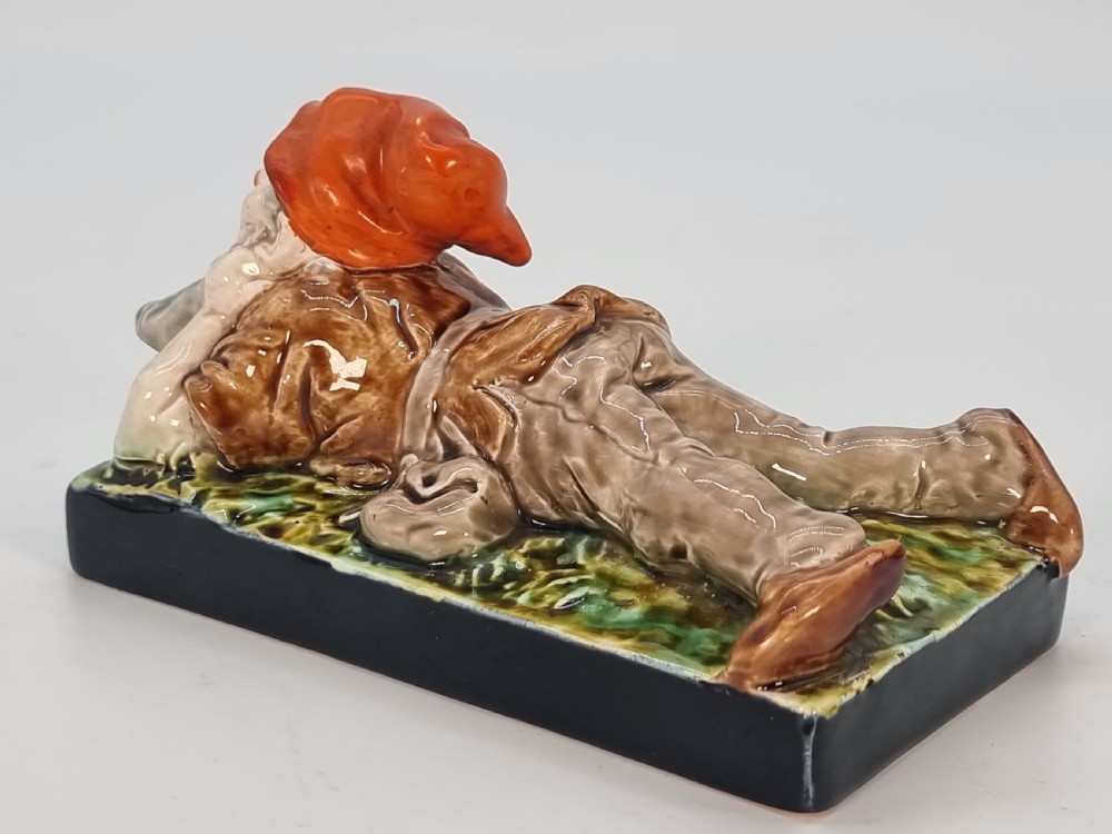 A Victorian majolica glazed earthenware paperweight depicting a reclining gnome with a red cap, grey beard, and brown tunic on a dark rectangular base. View V