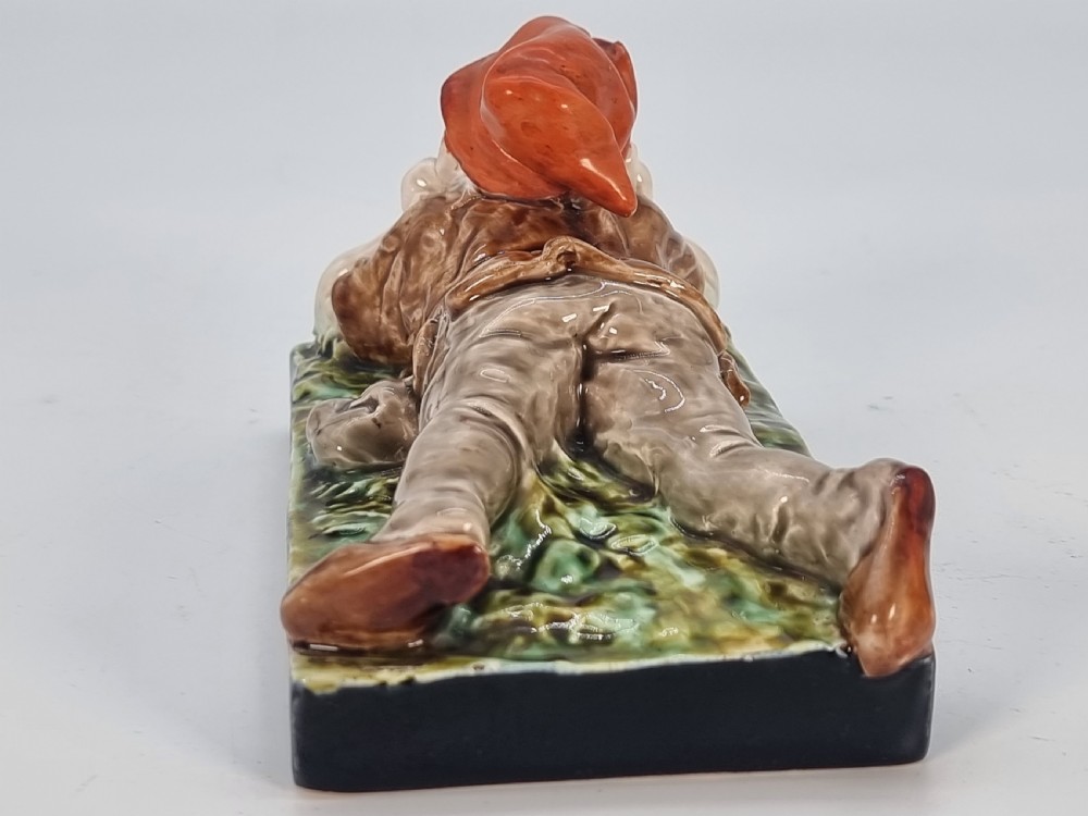 A Victorian majolica glazed earthenware paperweight depicting a reclining gnome with a red cap, grey beard, and brown tunic on a dark rectangular base. View VI