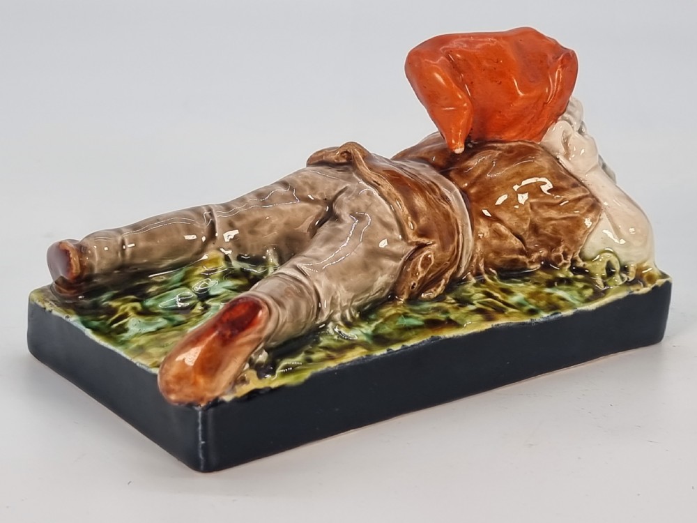 A Victorian majolica glazed earthenware paperweight depicting a reclining gnome with a red cap, grey beard, and brown tunic on a dark rectangular base. View VII