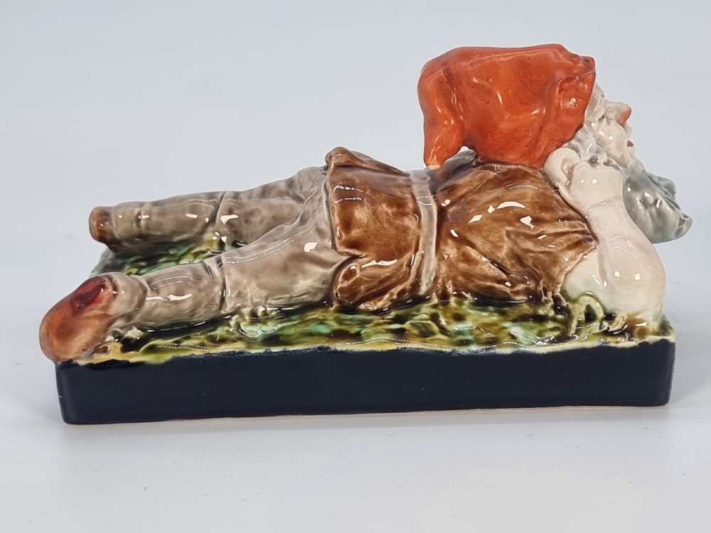 A Victorian majolica glazed earthenware paperweight depicting a reclining gnome with a red cap, grey beard, and brown tunic on a dark rectangular base. View VIII
