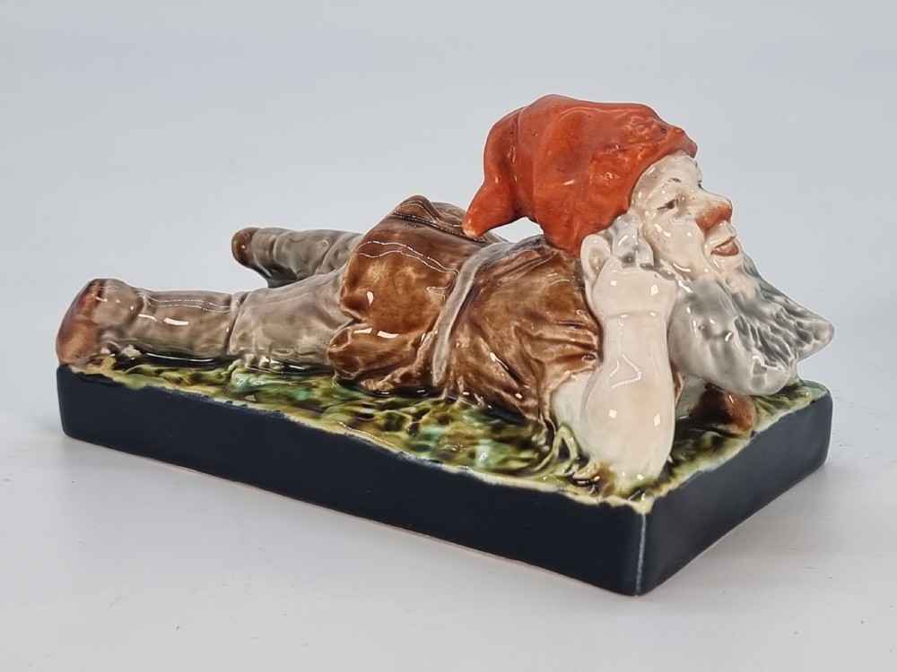 A Victorian majolica glazed earthenware paperweight depicting a reclining gnome with a red cap, grey beard, and brown tunic on a dark rectangular base. View IX