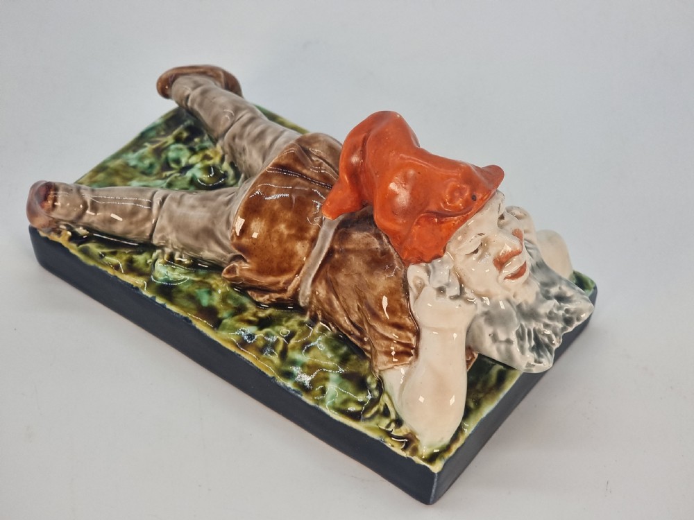 A Victorian majolica glazed earthenware paperweight depicting a reclining gnome with a red cap, grey beard, and brown tunic on a dark rectangular base. View X