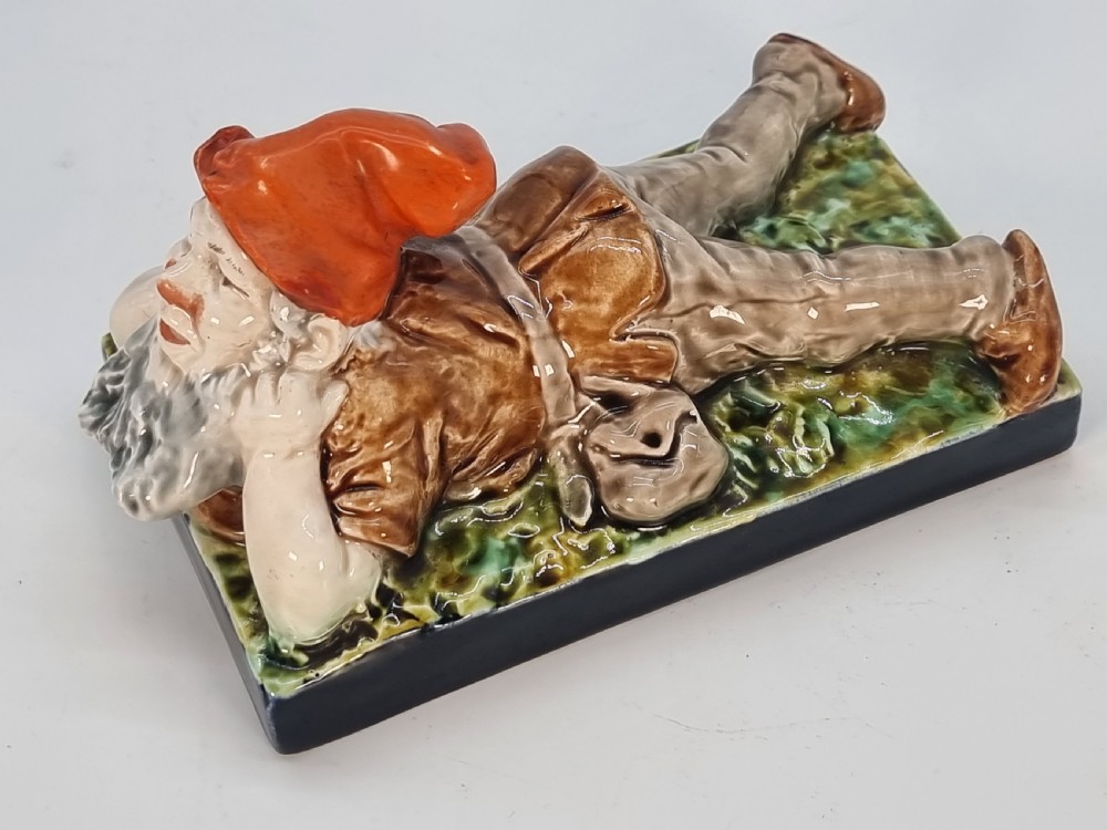 A Victorian majolica glazed earthenware paperweight depicting a reclining gnome with a red cap, grey beard, and brown tunic on a dark rectangular base. View XI