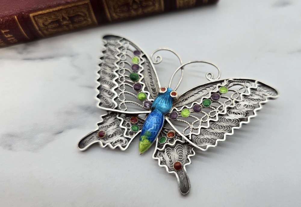 A silver filigree butterfly brooch featuring openwork wings filled with blue, green, purple, and red enamel details. View III