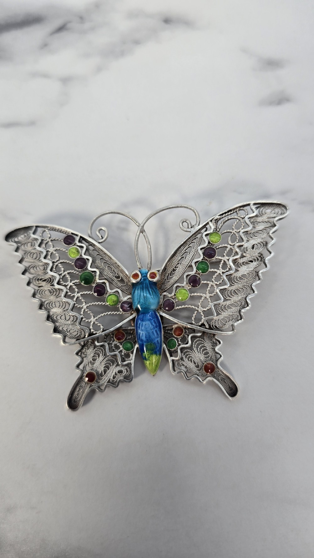 A silver filigree butterfly brooch featuring openwork wings filled with blue, green, purple, and red enamel details. View IV