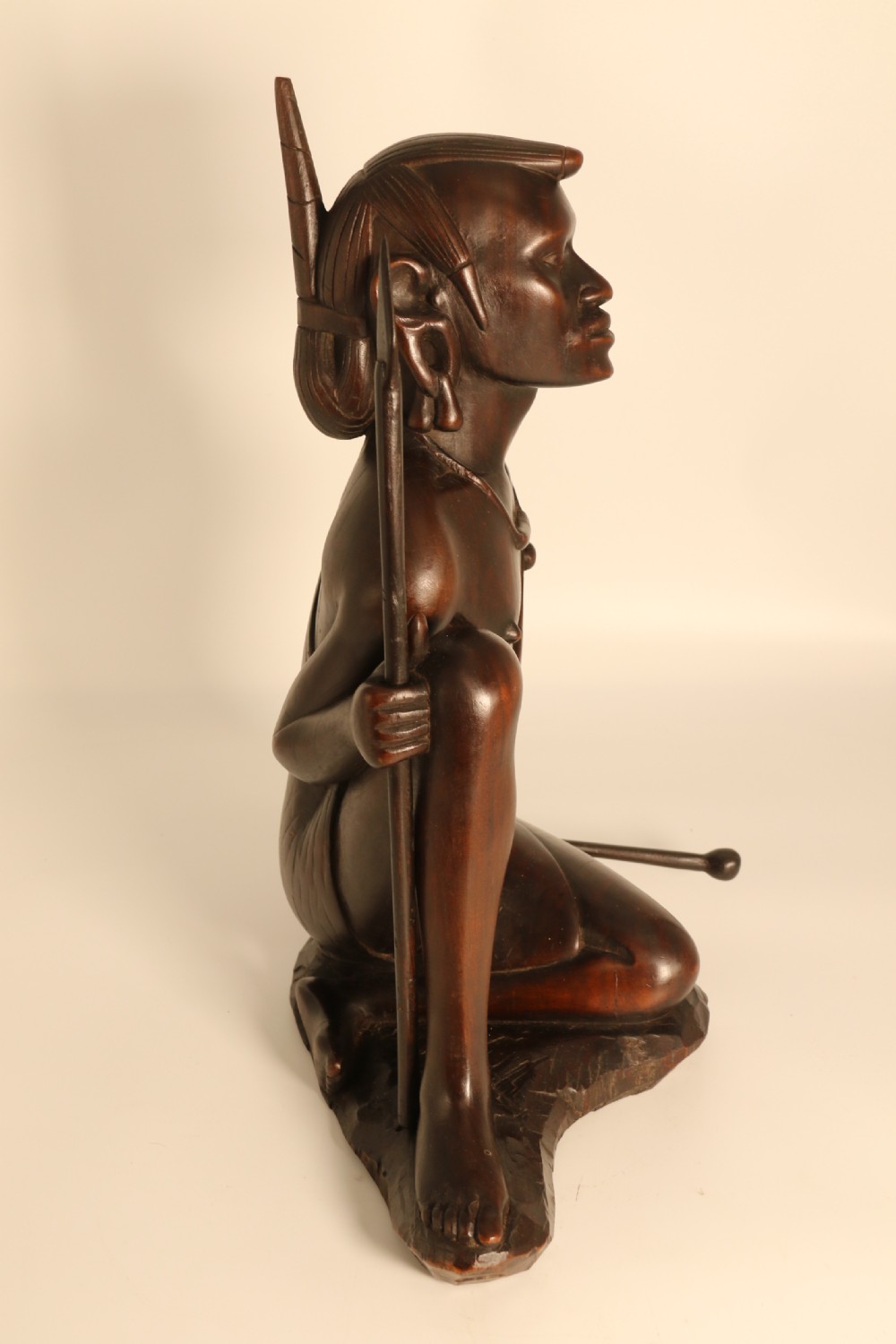A dark hardwood hand-carved sculpture of a kneeling Maasai warrior holding a spear and club on an integrated wooden base. View II