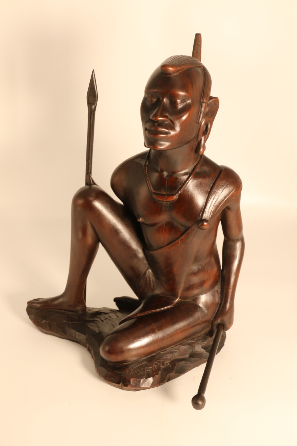 A dark hardwood hand-carved sculpture of a kneeling Maasai warrior holding a spear and club on an integrated wooden base. View X