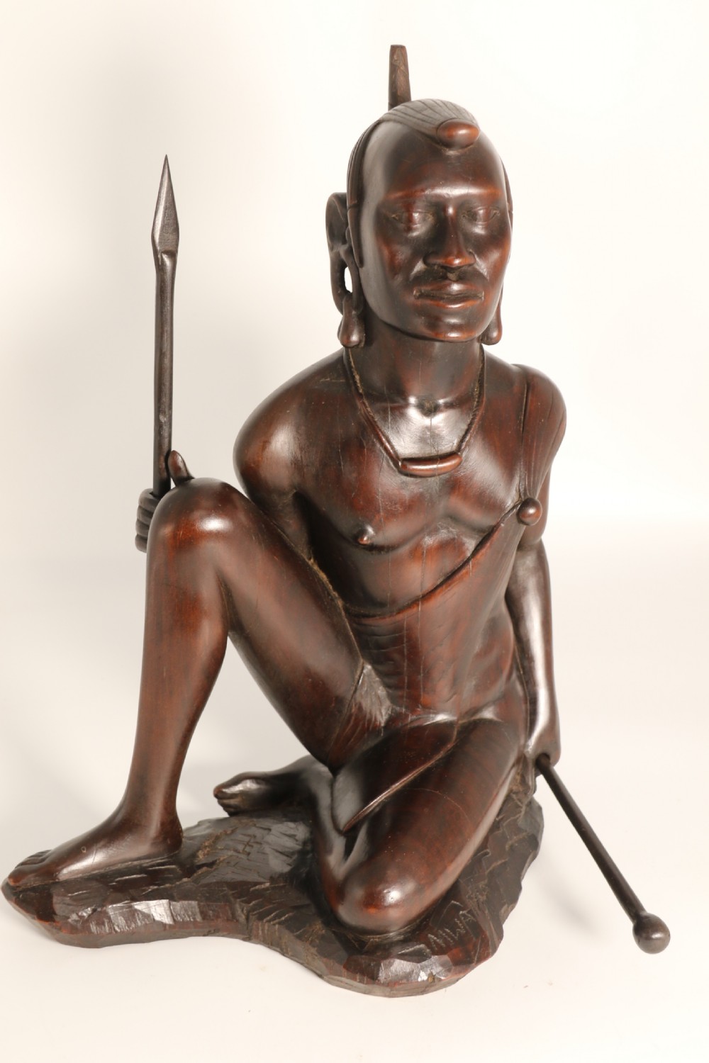 A dark hardwood hand-carved sculpture of a kneeling Maasai warrior holding a spear and club on an integrated wooden base.