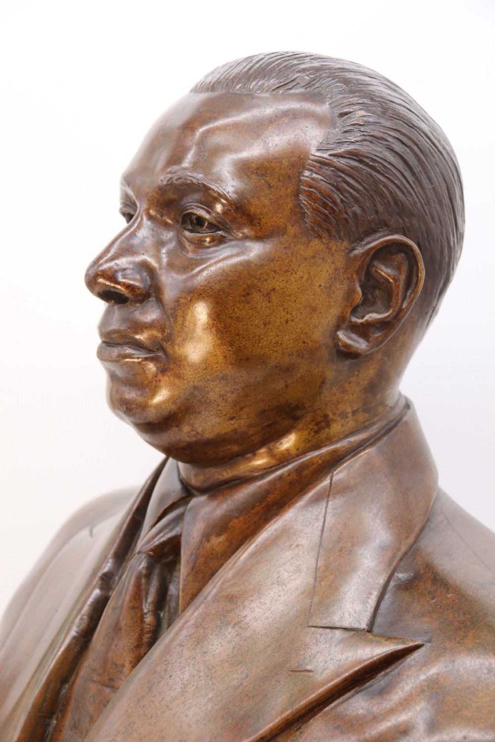 A cast bronze portrait bust depicting a gentleman in a suit jacket and tie, resting on a pedestal base with a square plinth. View IX
