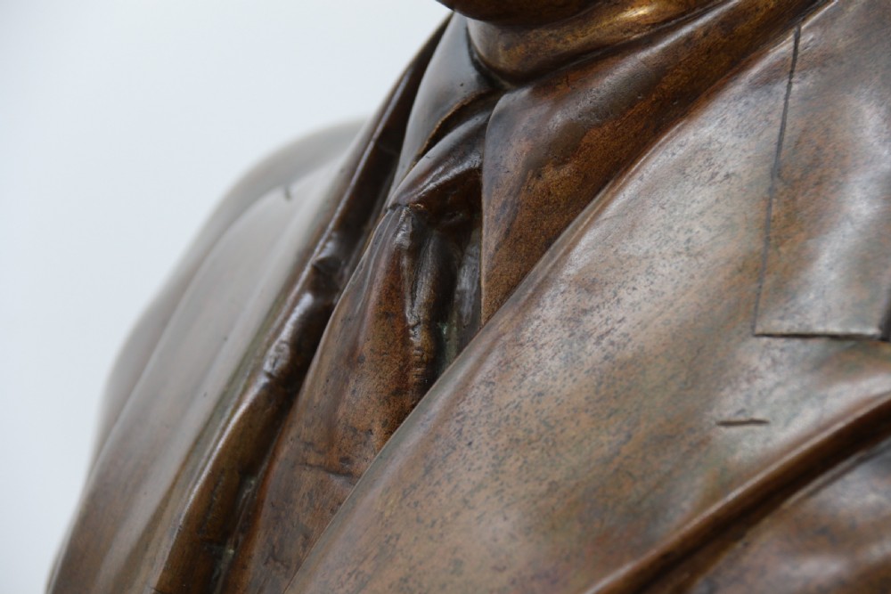 A cast bronze portrait bust depicting a gentleman in a suit jacket and tie, resting on a pedestal base with a square plinth. View X