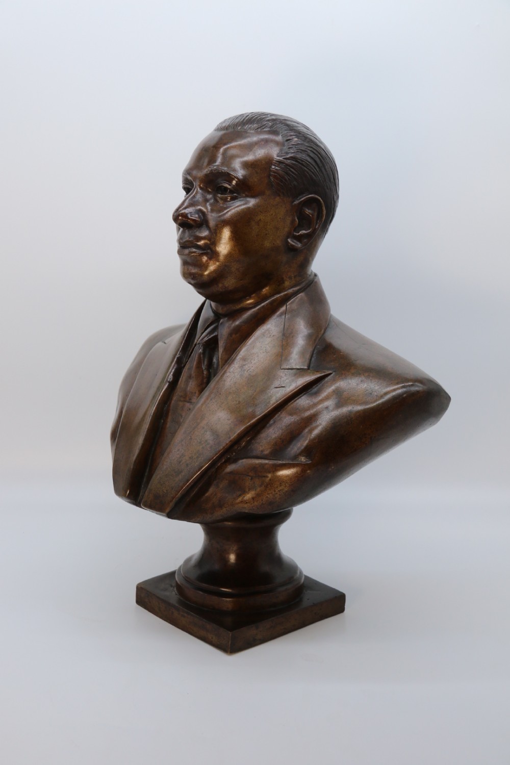 A cast bronze portrait bust depicting a gentleman in a suit jacket and tie, resting on a pedestal base with a square plinth. View XV