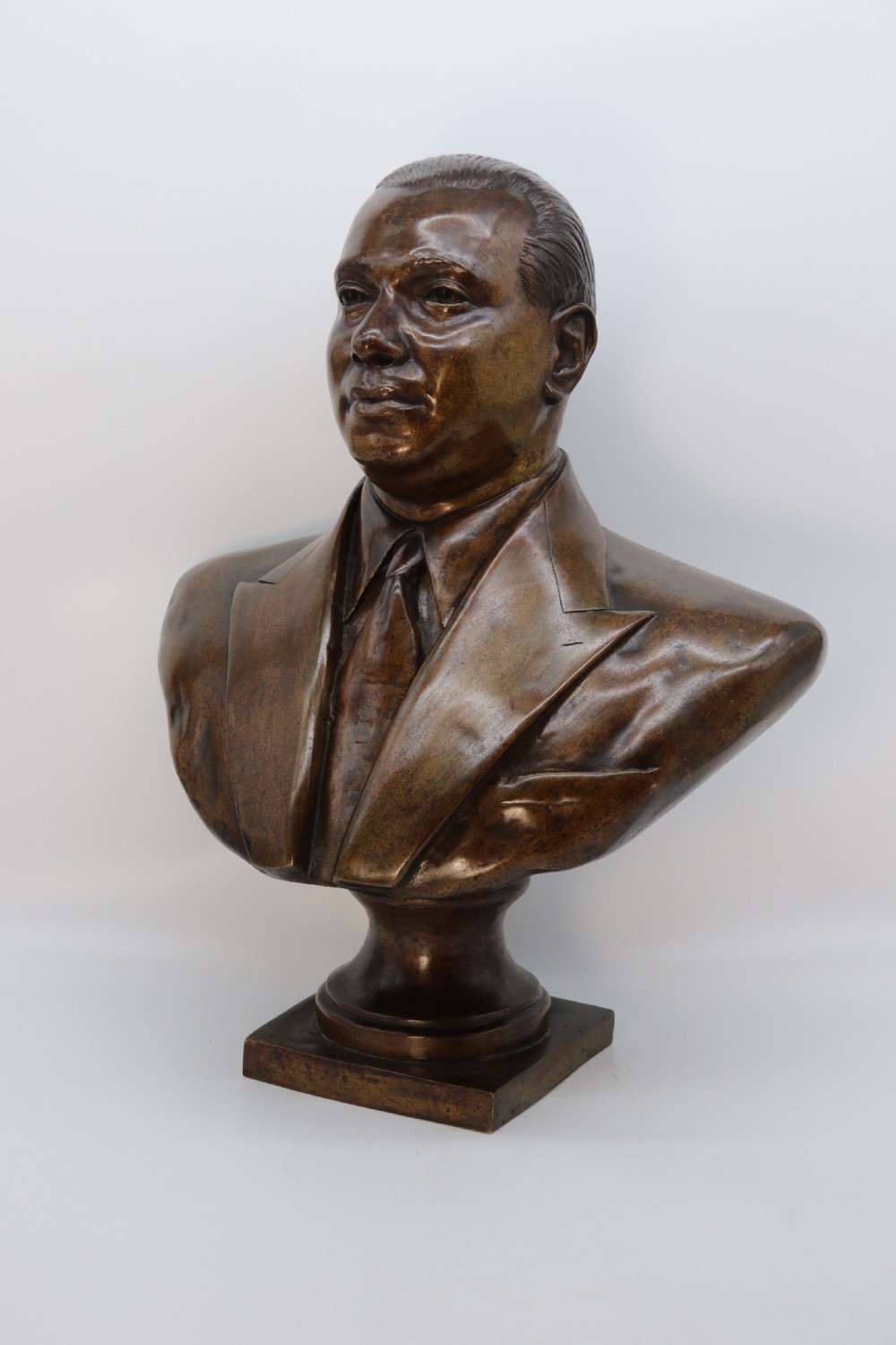 A cast bronze portrait bust depicting a gentleman in a suit jacket and tie, resting on a pedestal base with a square plinth. View XX