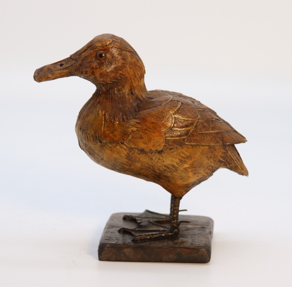 A small folk art duckling sculpture crafted from papier mache and textured straw, mounted on a square wooden base.
