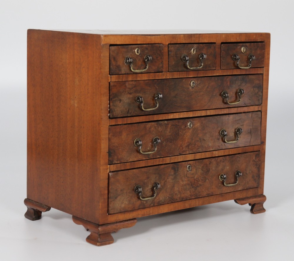 A miniature George II style chest of drawers crafted from figured walnut with brass drop handles, standing 20cm high beside a red apple for scale. View IV
