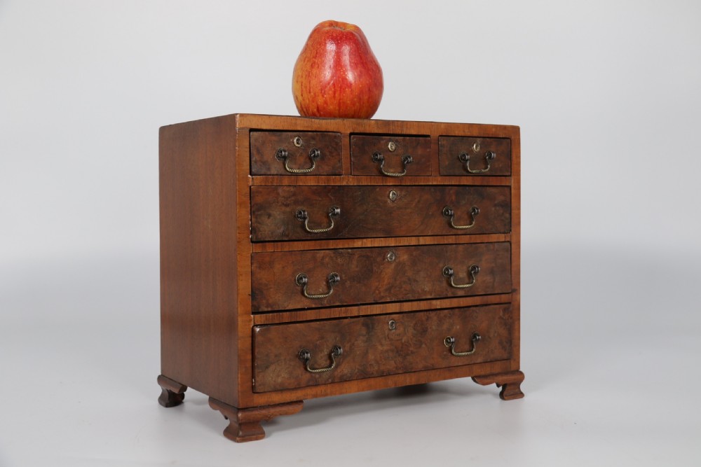 A miniature George II style chest of drawers crafted from figured walnut with brass drop handles, standing 20cm high beside a red apple for scale. View XIII