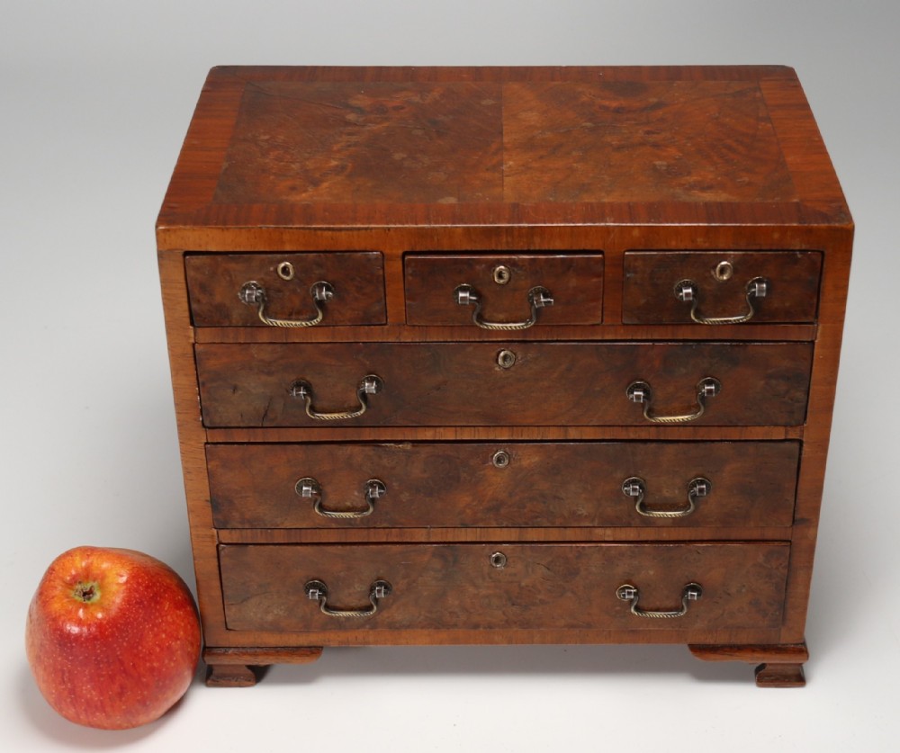 A miniature George II style chest of drawers crafted from figured walnut with brass drop handles, standing 20cm high beside a red apple for scale.