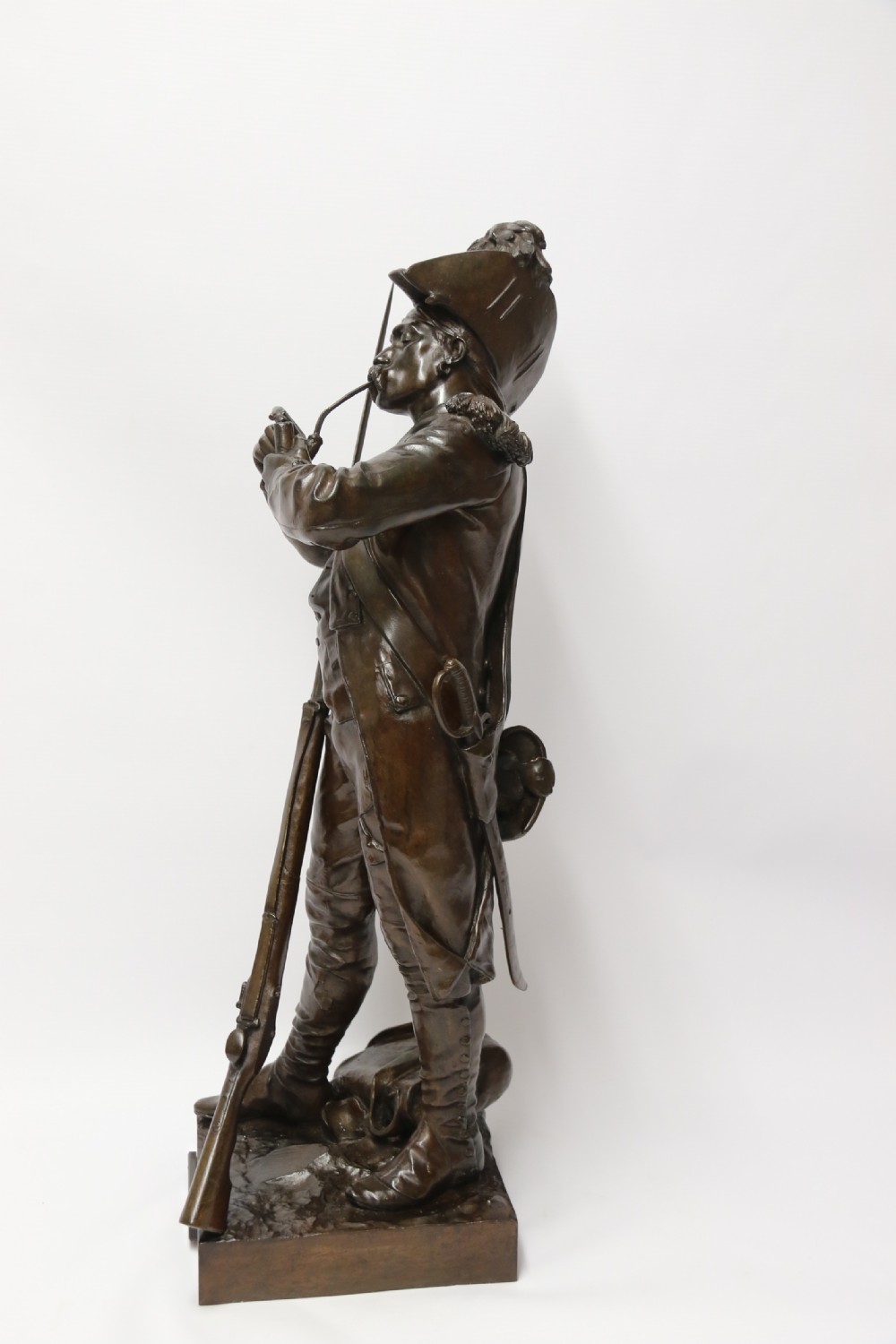 A dark brown patinated bronze sculpture depicting a 1792 French grenadier soldier in full uniform lighting a pipe beside a rifle. View II