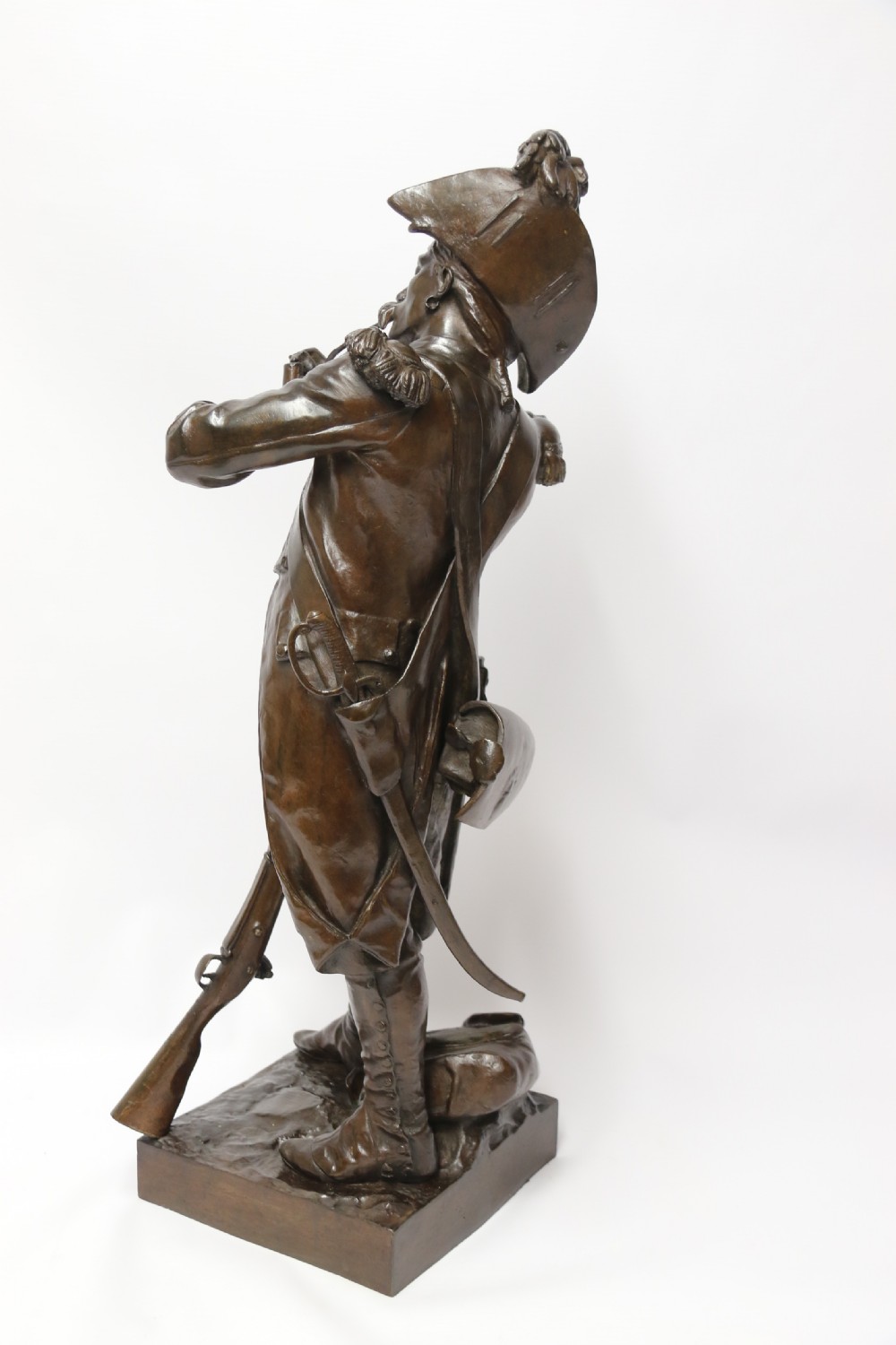 A dark brown patinated bronze sculpture depicting a 1792 French grenadier soldier in full uniform lighting a pipe beside a rifle. View III