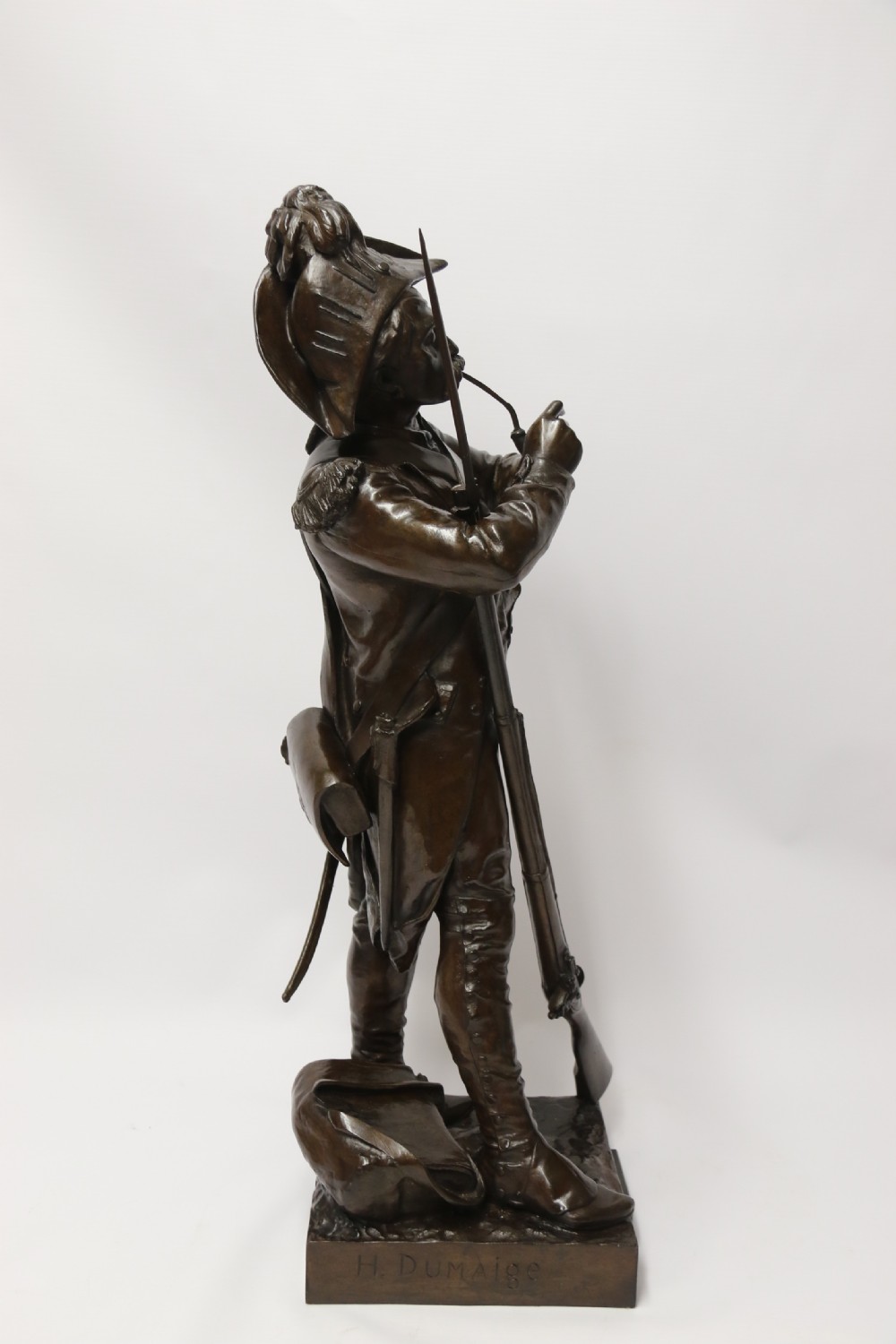 A dark brown patinated bronze sculpture depicting a 1792 French grenadier soldier in full uniform lighting a pipe beside a rifle. View IV