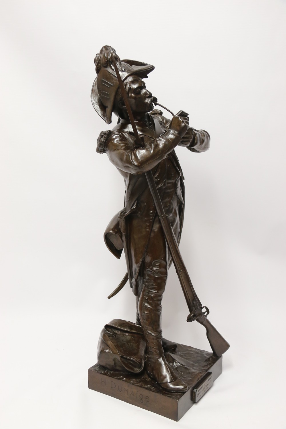 A dark brown patinated bronze sculpture depicting a 1792 French grenadier soldier in full uniform lighting a pipe beside a rifle. View VII