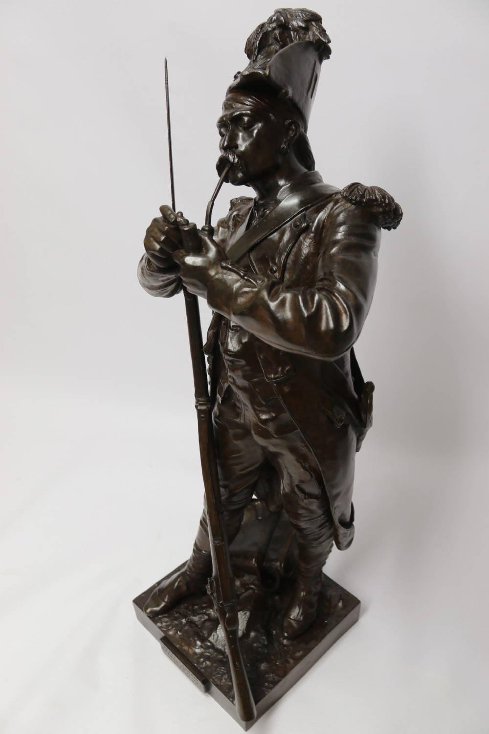 A dark brown patinated bronze sculpture depicting a 1792 French grenadier soldier in full uniform lighting a pipe beside a rifle. View XX