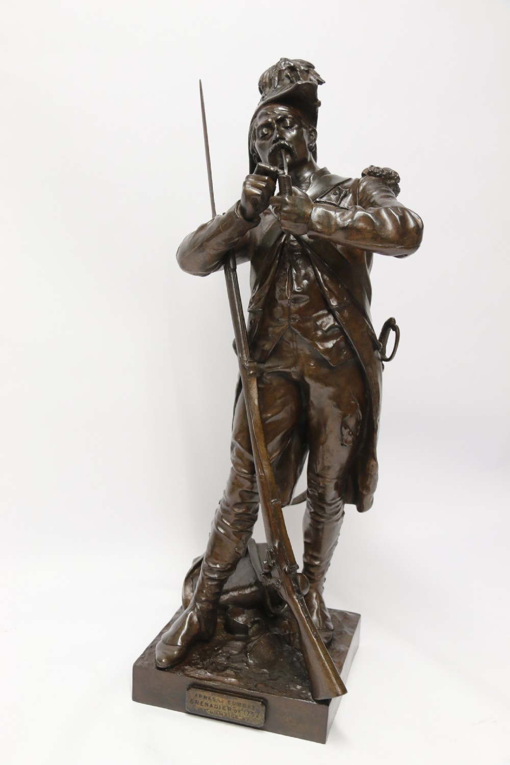 A dark brown patinated bronze sculpture depicting a 1792 French grenadier soldier in full uniform lighting a pipe beside a rifle. View XXI