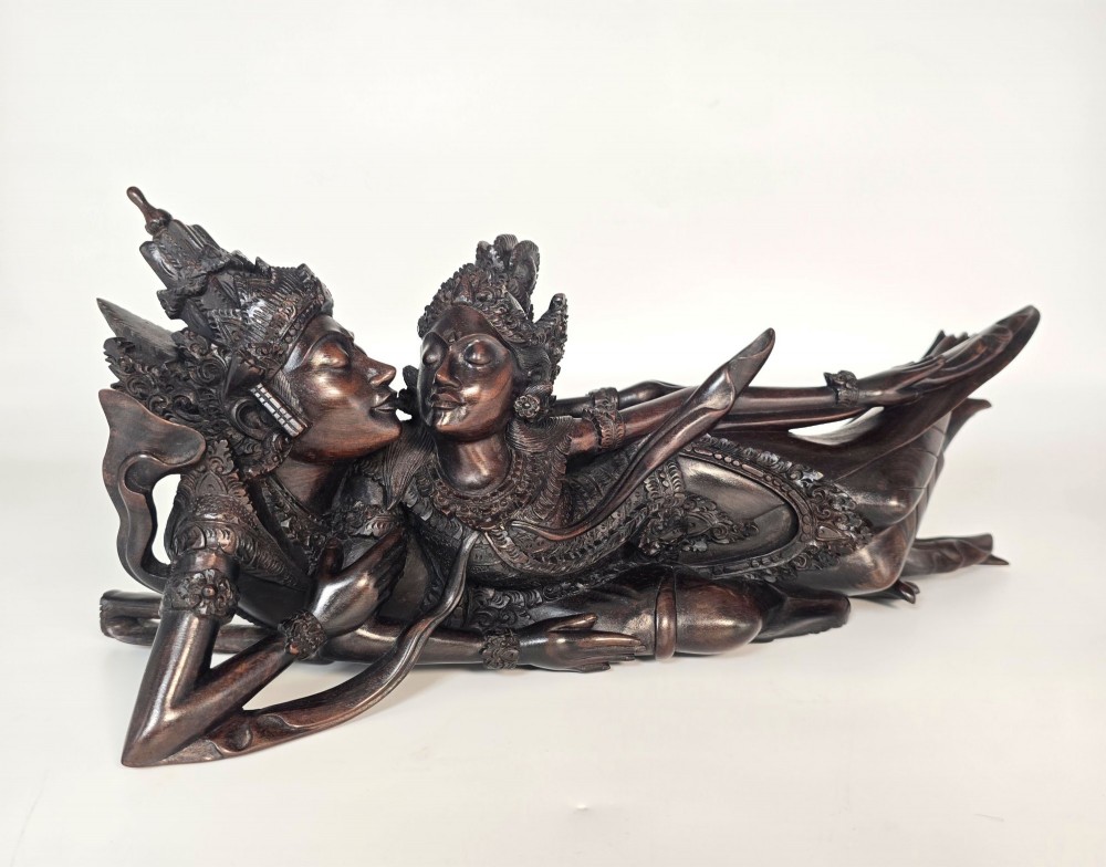 A dark hardwood Balinese carving depicting the reclining figures of Rama and Sita with detailed ornate headdresses and traditional attire.