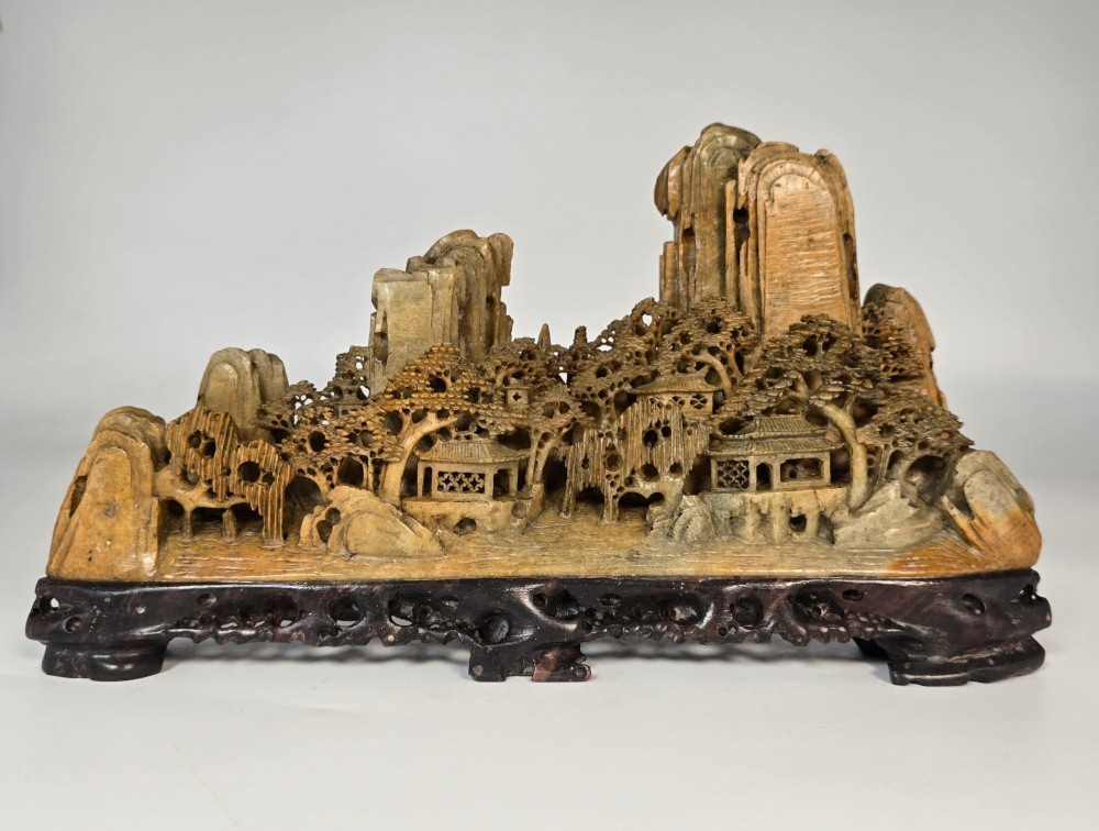 A carved Chinese soapstone continuous landscape featuring mountain peaks, pine trees, and pavilions set upon a fitted dark carved hardwood stand.