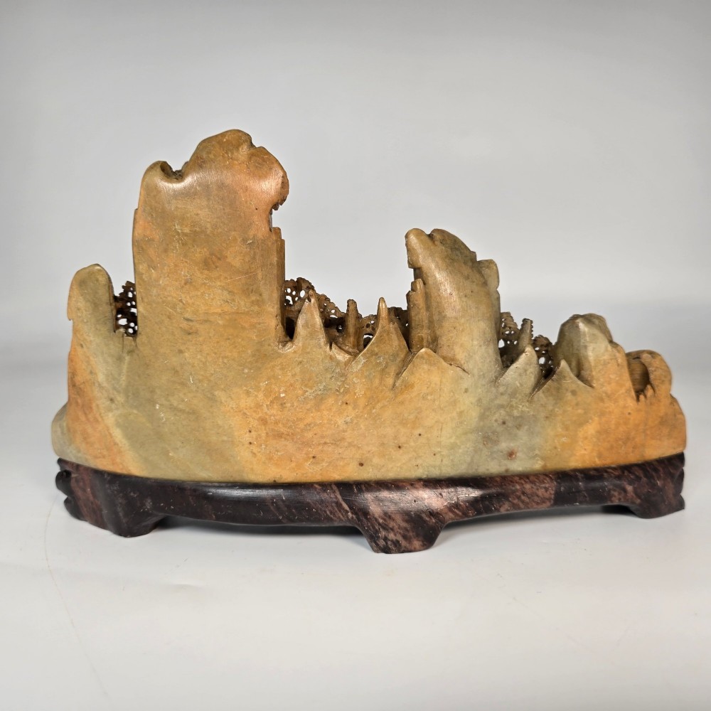 A carved Chinese soapstone continuous landscape featuring mountain peaks, pine trees, and pavilions set upon a fitted dark carved hardwood stand. View VI