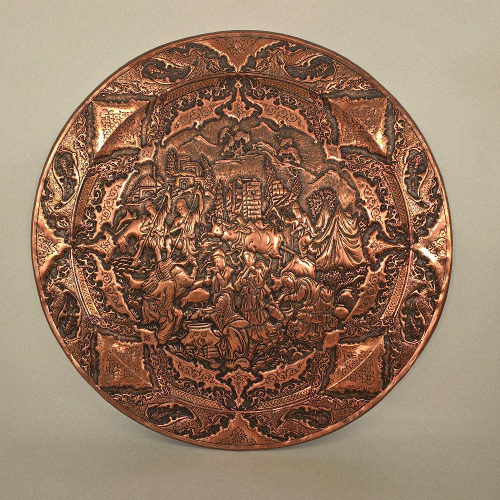 A circular Persian copper charger featuring detailed central repousse relief surrounded by intricate arabesque and paisley borders.