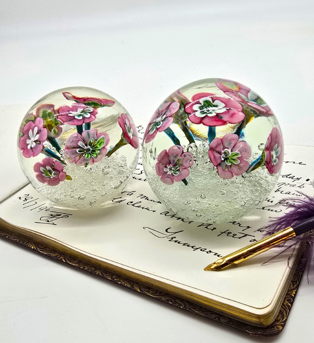 A pair of spherical clear glass paperweights containing pink lampwork flowers above a white bubble base layer.